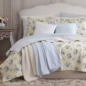 A bed set with a floral yellow comforter, blue throw, and decorative pillows on an elegant bed.