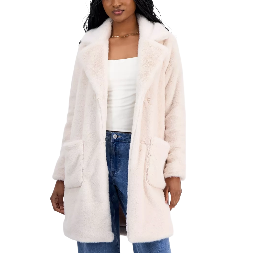 A woman is wearing a light beige faux fur coat with large front pockets, paired with a white top and blue jeans.