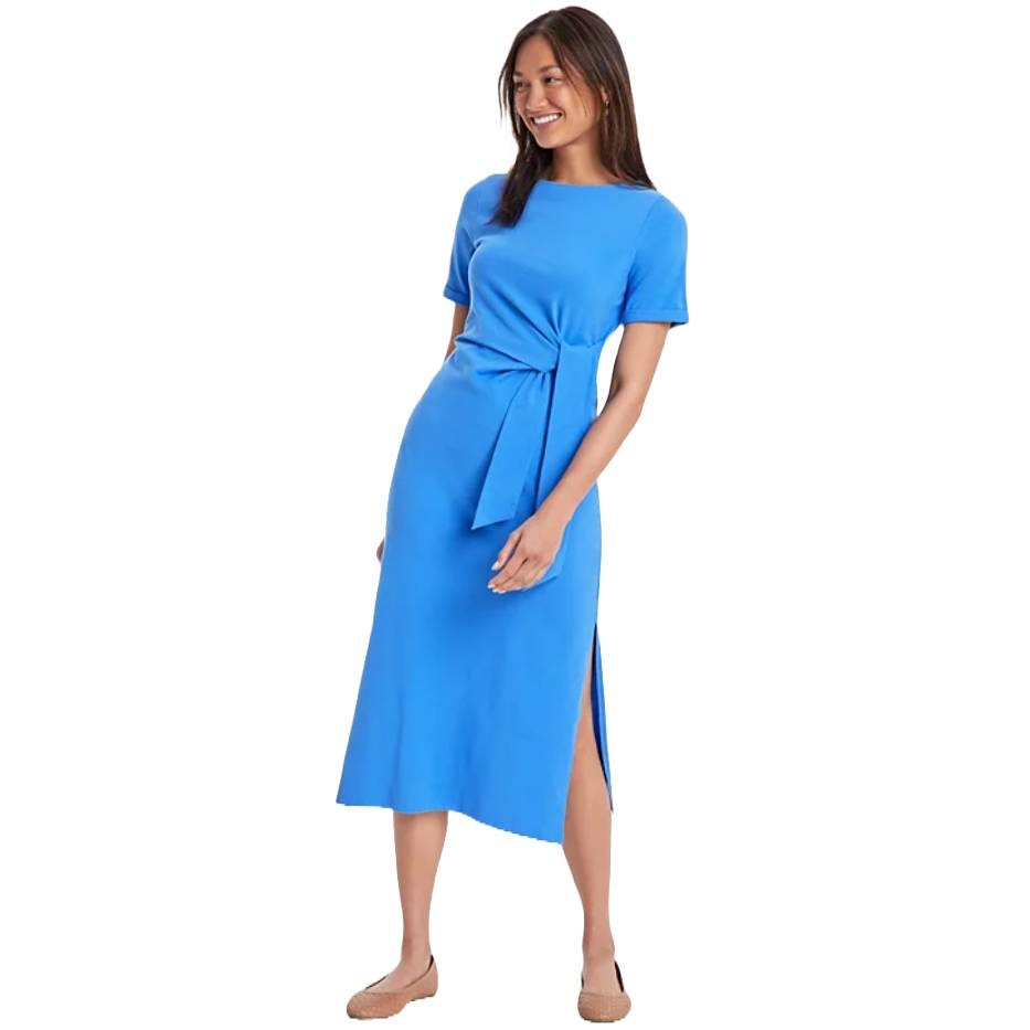 A woman is wearing a blue midi dress with short sleeves, a tie waist, and a side slit, paired with tan flats.