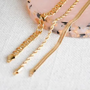 Three gold chains with varying link styles—curb, rope, and herringbone—are displayed on a light-colored textured surface next to a pink and black speckled background.