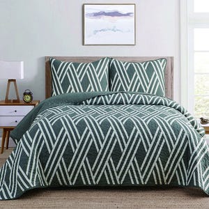 A bed is shown with a green and white geometric-patterned quilt and matching pillows, set in a modern bedroom with a nightstand, lamp, alarm clock, and wall art.