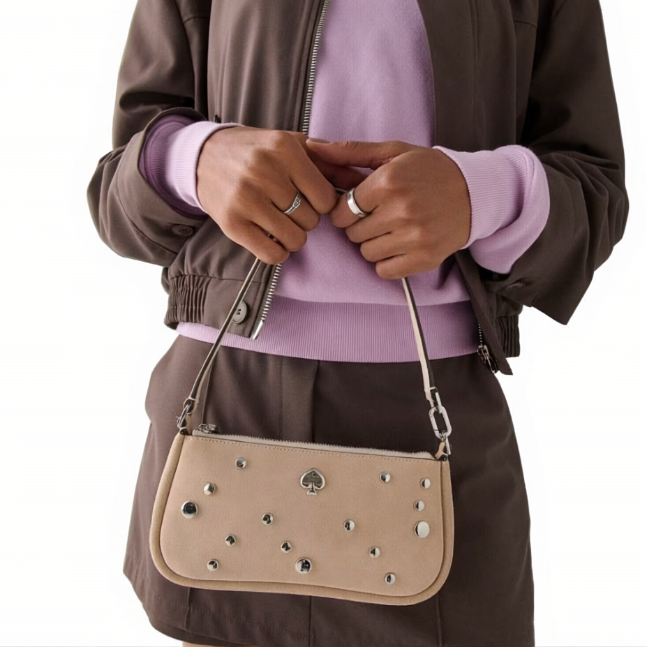 Person holding a beige handbag with silver studs and a spade logo, dressed in a brown jacket, matching skirt, and a lavender sweater.