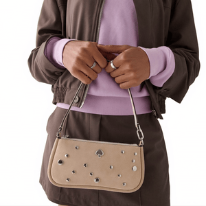 Person holding a beige handbag with silver studs and a spade logo, dressed in a brown jacket, matching skirt, and a lavender sweater.