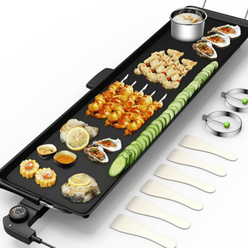 Electric griddle with various foods, including shrimp skewers, sliced cucumbers, and oysters. Accompanied by two metal rings, five spatulas, and visible controls for temperature adjustment.