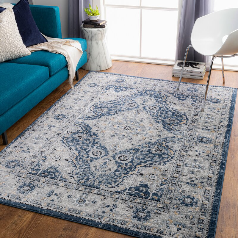 The 8' x 10' Kelly Clarkson Area Rug features a vintage floral pattern in shades of blue and gray, adding an elegant touch to the room. It lies on a wooden floor beside a teal sofa and a white chair, with natural light streaming in from a window.