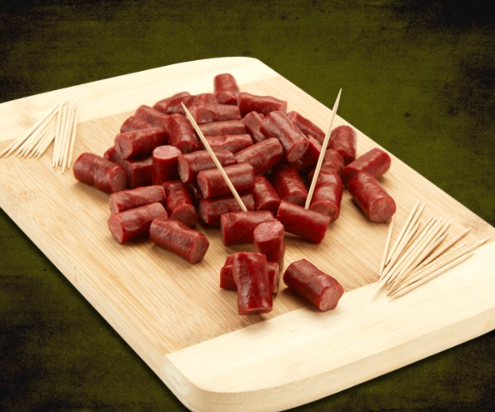 A wooden board displays numerous bite-sized beef or turkey snack sticks, dark red in color, alongside scattered wooden toothpicks for serving.