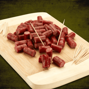 A wooden board displays numerous bite-sized beef or turkey snack sticks, dark red in color, alongside scattered wooden toothpicks for serving.