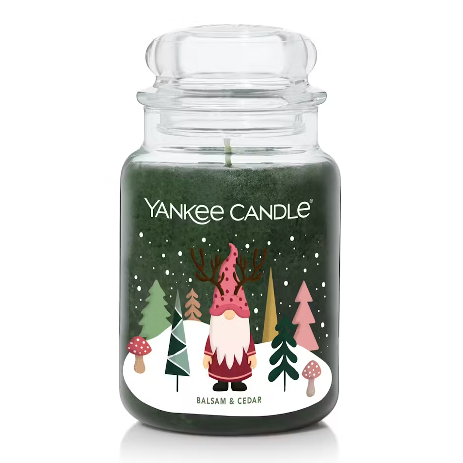 A Yankee Candle in a large jar with a Balsam & Cedar scent features a festive design, including a gnome with antlers surrounded by stylized trees and mushrooms on a snowy background.