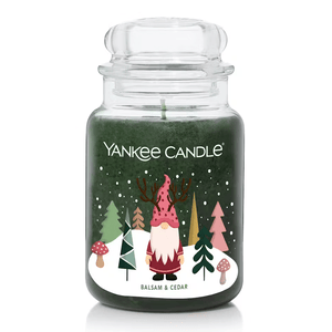 A Yankee Candle in a large jar with a Balsam & Cedar scent features a festive design, including a gnome with antlers surrounded by stylized trees and mushrooms on a snowy background.