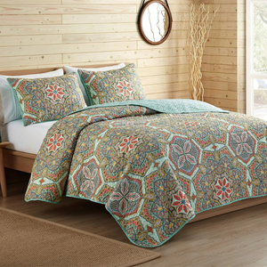 A bed set with a colorful, intricate paisley and floral pattern on a quilt and matching pillow shams, placed in a wooden-paneled room with a round mirror and decorative branches.