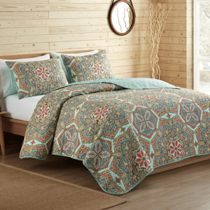 A bed set with a colorful, intricate paisley and floral pattern on a quilt and matching pillow shams, placed in a wooden-paneled room with a round mirror and decorative branches.
