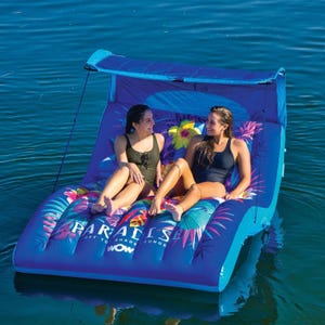The Wow Sports Paradise Lounger is a blue inflatable two-seater raft with a colorful tropical design and a detachable sunshade, designed for relaxing on the water. Two people are shown enjoying its comfortable surface.