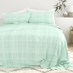 A bed with a light green, plaid-patterned bedspread and matching pillows in a minimalist setting, featuring white and plant accents.