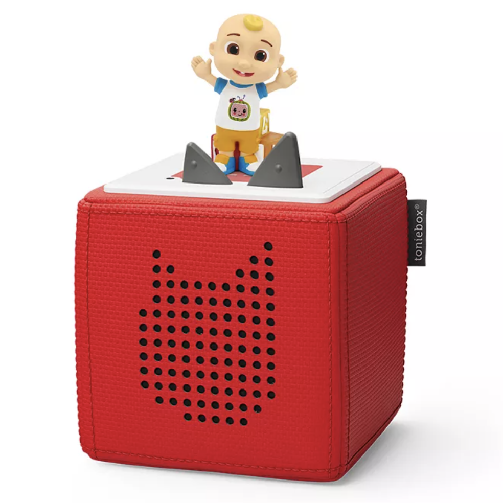 A red Toniebox with a perforated speaker design features a figurine of a smiling baby on top, used for playing audio content, such as stories, when figurines are placed on it.