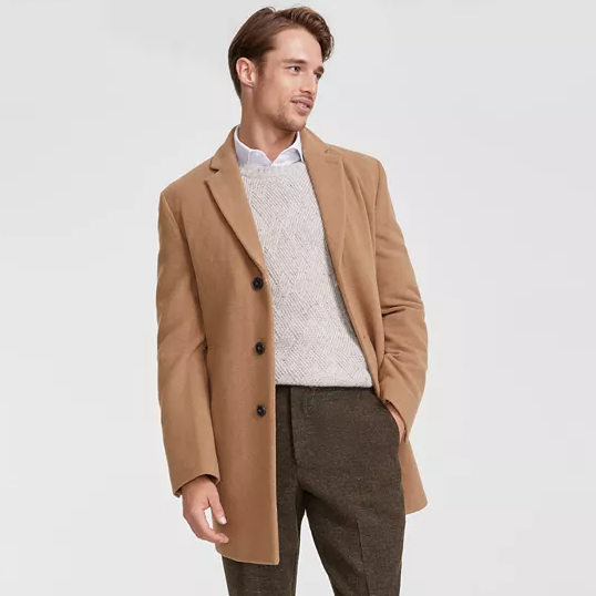 A man is wearing a camel-colored Calvin Klein wool-blend overcoat, featuring a notch lapel, button front, and side pockets, styled with a cable-knit sweater and wool trousers.