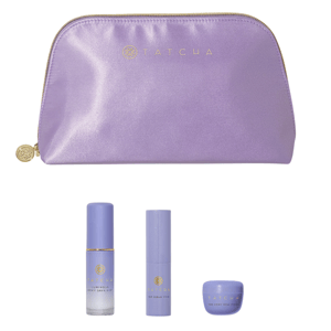 A lavender Tatcha pouch contains three skincare items: Luminous Dewy Skin Mist, The Serum Stick, and The Dewy Skin Cream, all in matching purple packaging.