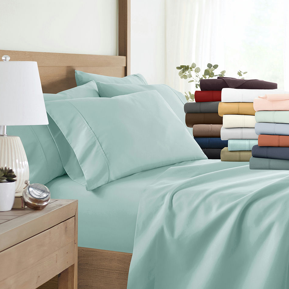 A neatly made bed with light blue sheets and pillowcases is displayed beside a stack of folded sheets in various colors, including red, brown, white, and peach. A bedside table with a lamp and a small plant is also visible.