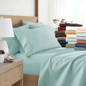 A neatly made bed with light blue sheets and pillowcases is displayed beside a stack of folded sheets in various colors, including red, brown, white, and peach. A bedside table with a lamp and a small plant is also visible.