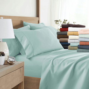 A neatly made bed with light blue sheets and pillowcases is displayed beside a stack of folded sheets in various colors, including red, brown, white, and peach. A bedside table with a lamp and a small plant is also visible.