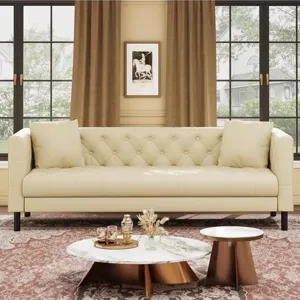 A cream-colored tufted sofa with cushions is in front of large windows with brown curtains. A round marble coffee table and a smaller side table are on a patterned rug, featuring a decorative tray with glassware and a vase.