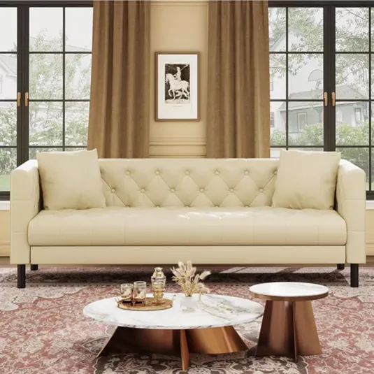 A cream-colored tufted sofa with cushions is in front of large windows with brown curtains. A round marble coffee table and a smaller side table are on a patterned rug, featuring a decorative tray with glassware and a vase.