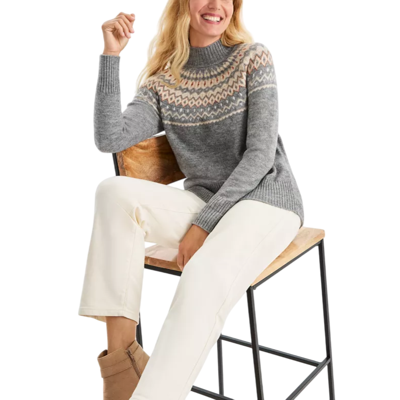 A woman is wearing a gray knit sweater with a colorful geometric pattern across the chest and shoulders, paired with white pants and tan ankle boots, sitting on a wooden chair with a black metal frame.