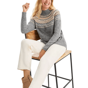 A woman is wearing a gray knit sweater with a colorful geometric pattern across the chest and shoulders, paired with white pants and tan ankle boots, sitting on a wooden chair with a black metal frame.