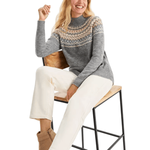 A woman is wearing a gray knit sweater with a colorful geometric pattern across the chest and shoulders, paired with white pants and tan ankle boots, sitting on a wooden chair with a black metal frame.
