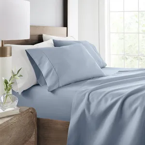 Light blue bedsheet and pillowcases on a wooden bed, accompanied by white pillows, near a window with natural light. A wooden bedside table features a lamp, a small plant, and a book.