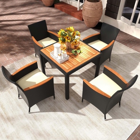 A 5-piece patio dining set featuring a square table with a wooden top and four cushioned chairs with dark wicker frames and wooden armrests. A vase with sunflowers is placed on the table. The set is positioned on a light-colored outdoor rug.