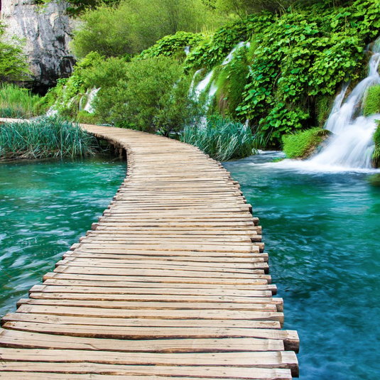 A wooden boardwalk curves through a lush, green landscape, crossing over vibrant turquoise water with small waterfalls and rich foliage surrounding it.