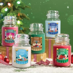 Five Yankee Candle jars in festive scents: Jolly Berry, North Pole Frost, VYuletide Wishes, Gingerbread Nutcrackers, and Big Apple Christmas are displayed with a holiday backdrop featuring a Christmas tree and wrapped gifts.