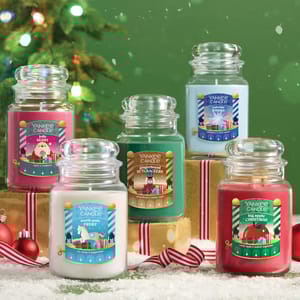 Five Yankee Candle jars in festive scents: Jolly Berry, North Pole Frost, VYuletide Wishes, Gingerbread Nutcrackers, and Big Apple Christmas are displayed with a holiday backdrop featuring a Christmas tree and wrapped gifts.