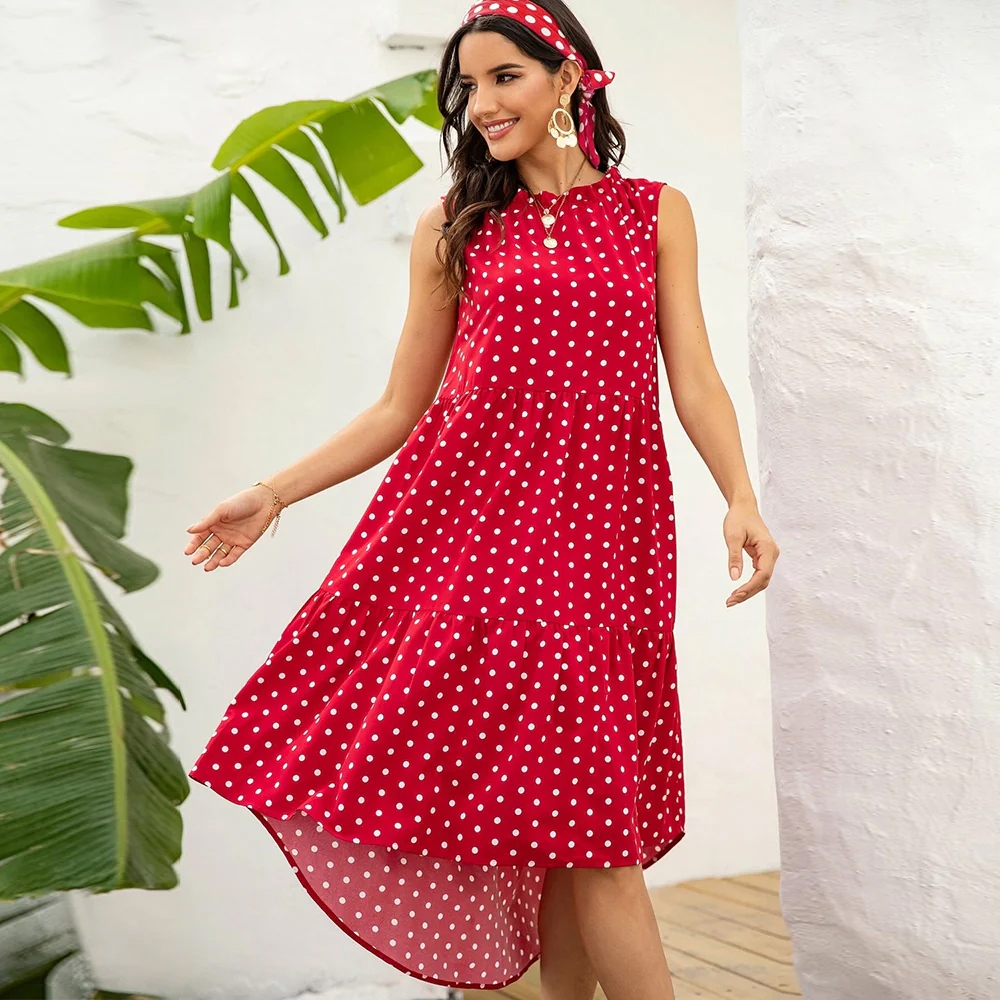 A woman is wearing a sleeveless red dress with white polka dots, paired with a matching headband. She is outdoors near green plants and a white wall, smiling and appearing relaxed.
