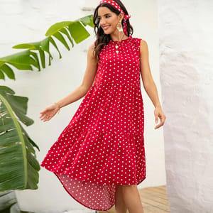 A woman is wearing a sleeveless red dress with white polka dots, paired with a matching headband. She is outdoors near green plants and a white wall, smiling and appearing relaxed.