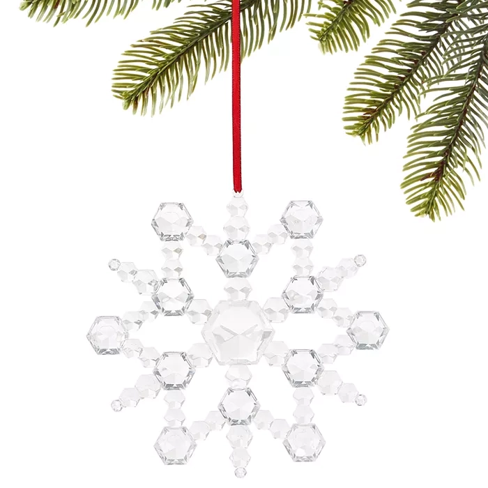 A faceted crystal snowflake ornament hangs from a red ribbon. It's delicately constructed with hexagonal shapes, reflecting light, and is displayed against green pine branches.