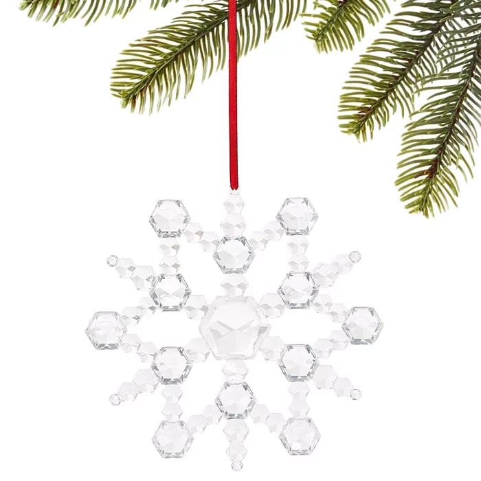 A faceted crystal snowflake ornament hangs from a red ribbon. It's delicately constructed with hexagonal shapes, reflecting light, and is displayed against green pine branches.