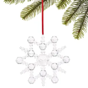 A faceted crystal snowflake ornament hangs from a red ribbon. It's delicately constructed with hexagonal shapes, reflecting light, and is displayed against green pine branches.
