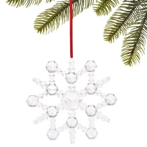 A faceted crystal snowflake ornament hangs from a red ribbon. It's delicately constructed with hexagonal shapes, reflecting light, and is displayed against green pine branches.