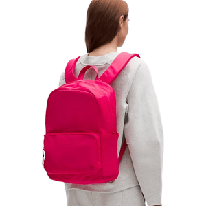 A person with long hair is wearing a vibrant pink backpack, featuring padded straps and a front pocket, paired with a light gray outfit.