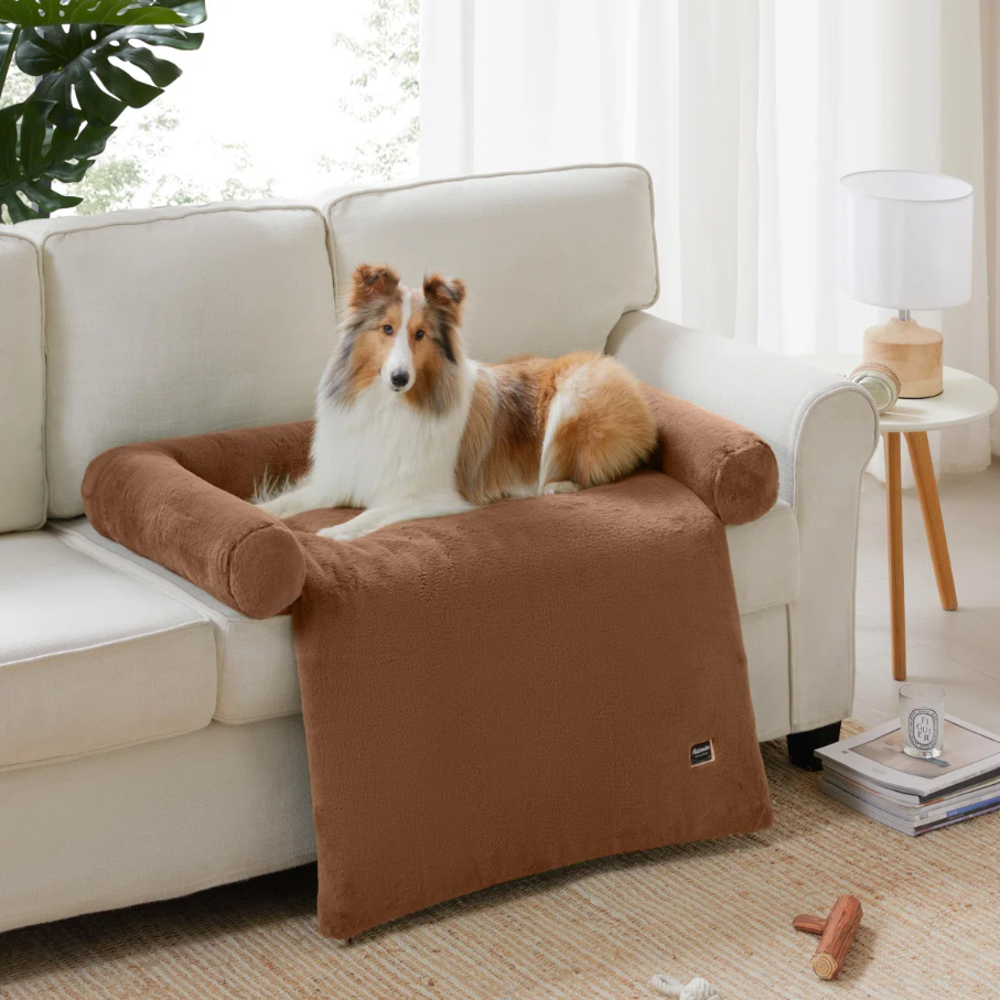 A dog is lying on a brown, plush, couch armrest cover that protects the furniture, offering a comfortable space. A side table with a lamp and a few small items are nearby.
