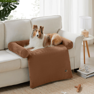 A dog is lying on a brown, plush, couch armrest cover that protects the furniture, offering a comfortable space. A side table with a lamp and a few small items are nearby.