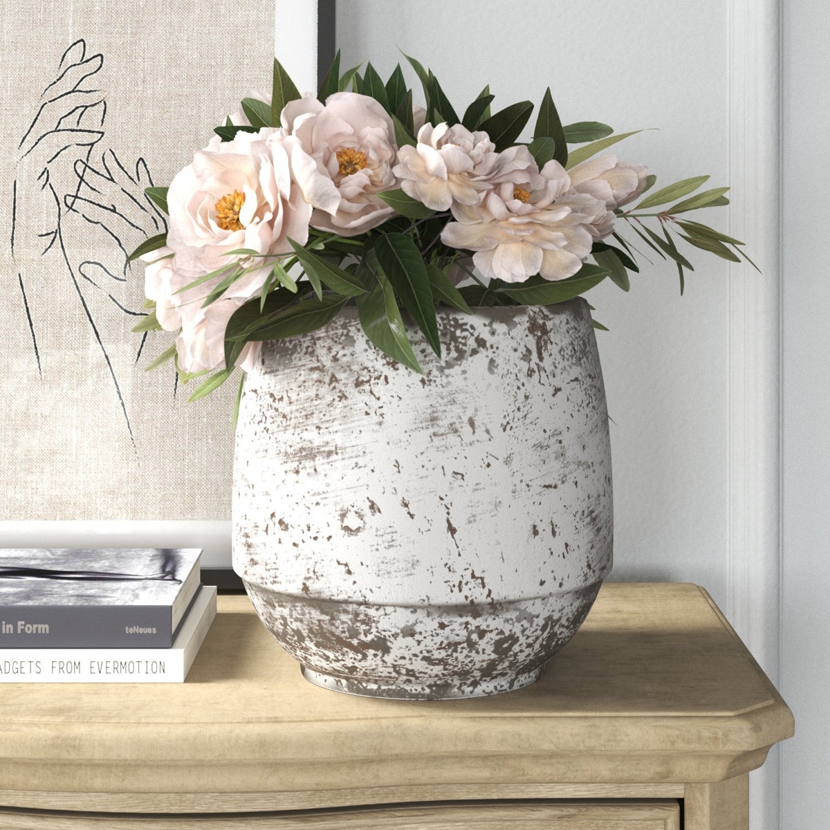 A rustic white vase with a distressed finish holds pink peonies and greenery, placed on a light wood table beside a line art print of hands and a stack of books.