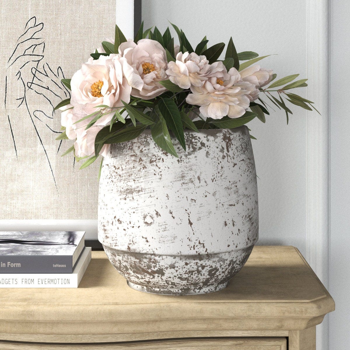 A rustic white vase with a distressed finish holds pink peonies and greenery, placed on a light wood table beside a line art print of hands and a stack of books.