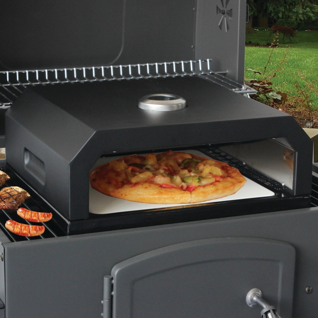 Outdoor pizza oven attachment on a barbecue grill with a pizza being baked inside and sausages and grilled vegetables on the side.