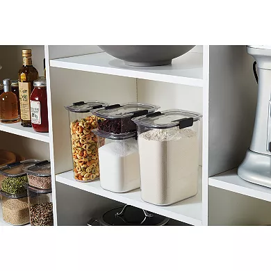 Eight clear Rubbermaid Brilliance containers of varying sizes with black latching lids, used for food storage, are displayed on kitchen shelves.
