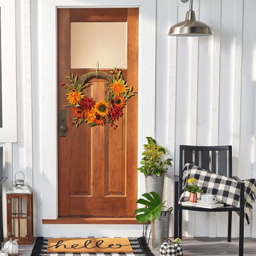 A front porch features a wooden door with a fall-themed wreath, a \“hello\“ doormat, a black chair with a plaid cushion, a small table with a cup, potted plants, and a lantern.