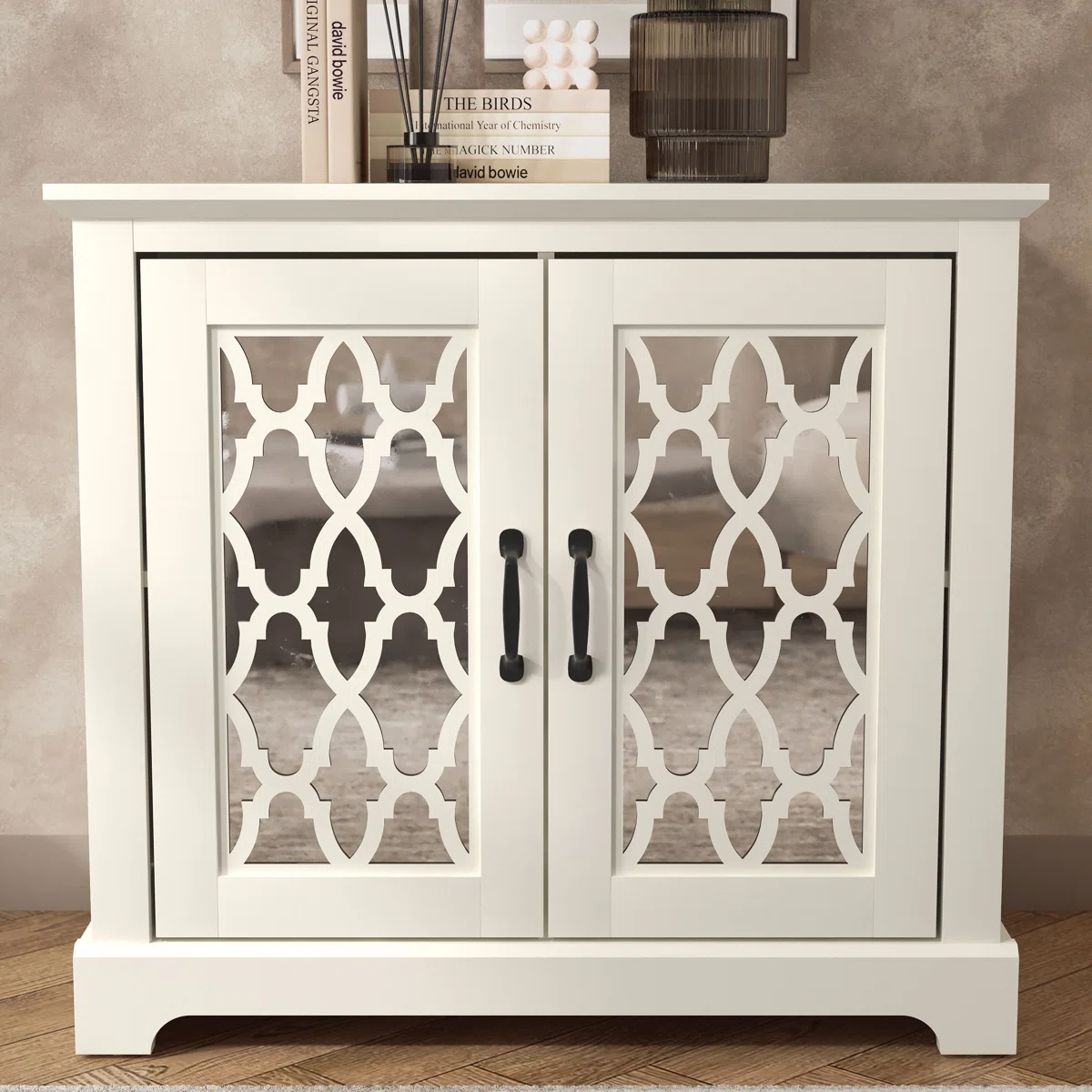 White cabinet with decorative mirrored doors, featuring a geometric pattern and black handles. Two books, an ornament, and a dark glass are placed on top.