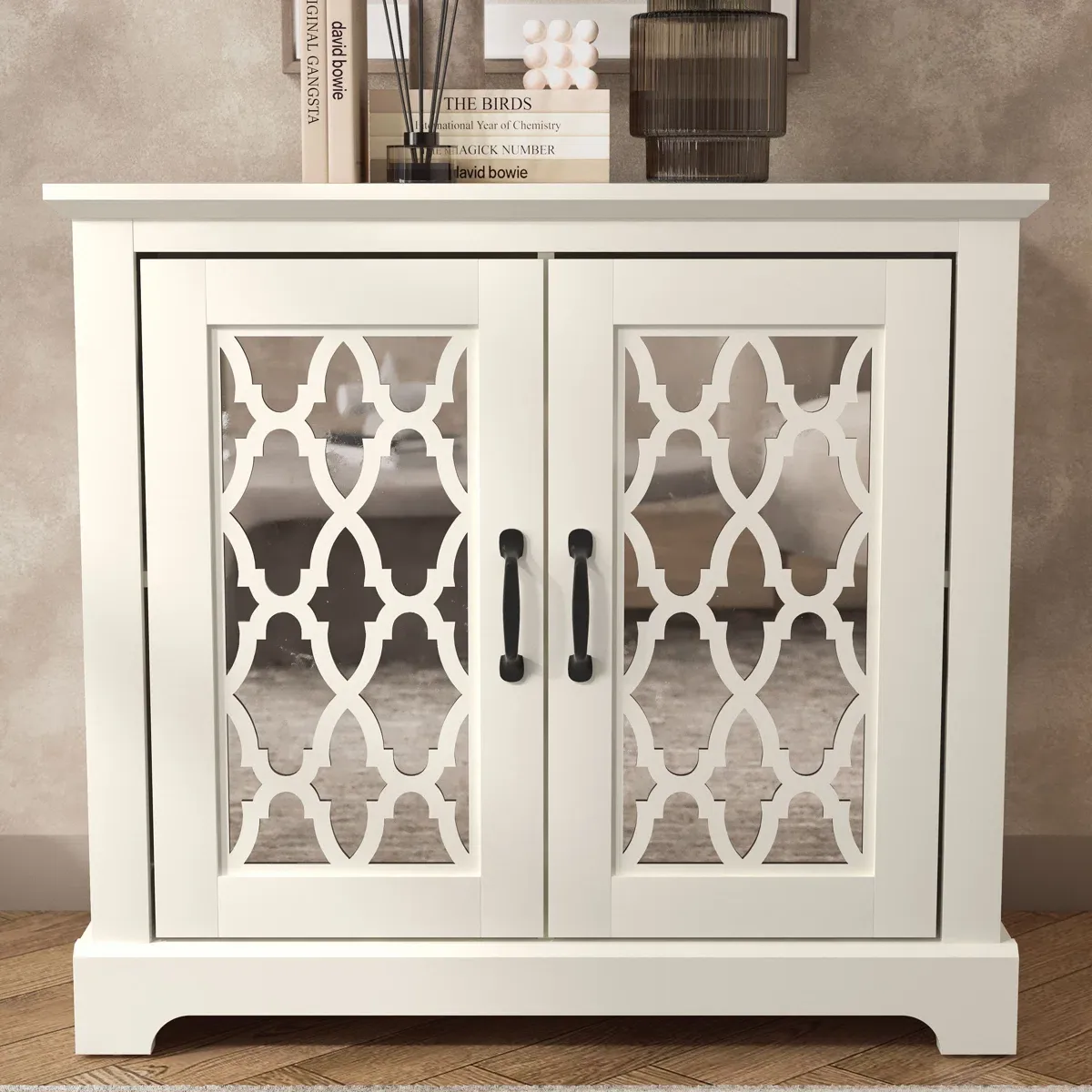White cabinet with decorative mirrored doors, featuring a geometric pattern and black handles. Two books, an ornament, and a dark glass are placed on top.