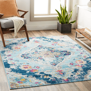 A 5' x 7' area rug features a vibrant medallion design with shades of blue, pink, and cream. It's placed on light wooden flooring, accompanied by a mid-century modern chair, a planter, and a table with books.